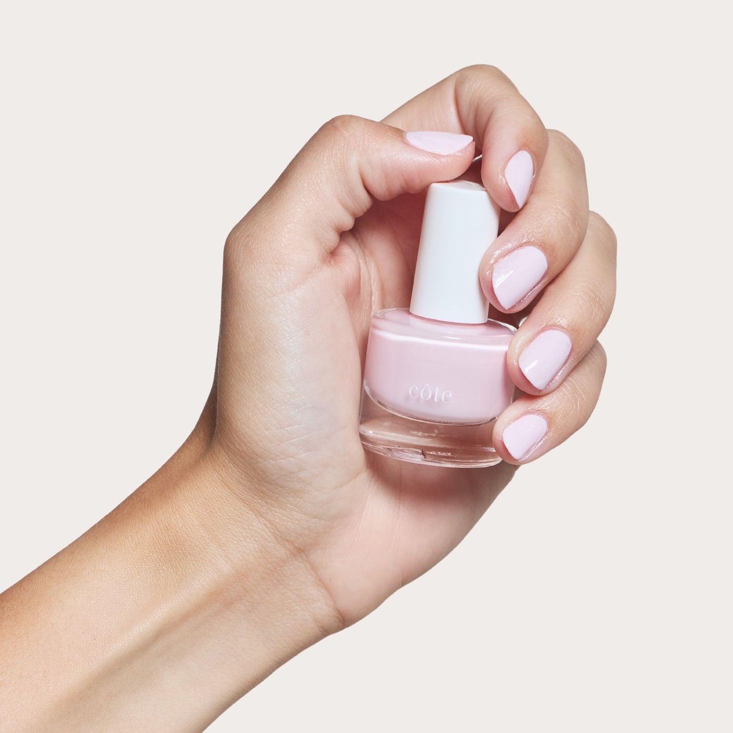 Hand holding Côte Beauty Moonbeam Soft Blush Pink Non-Toxic Nail Polish bottle with nails painted. 