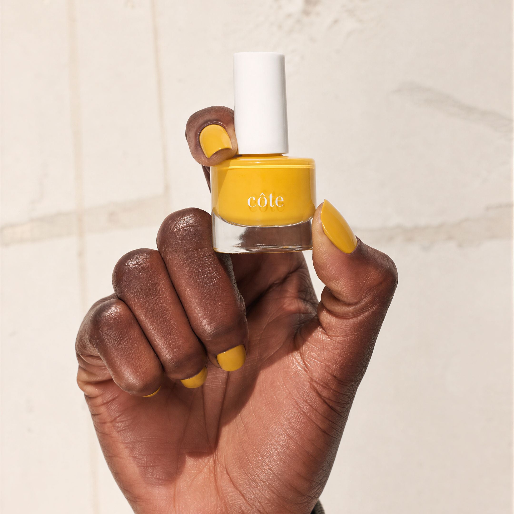 Hand holding bottle of nail polish with nails painted with Côte Beauty Songbird dandelion yellow non-toxic nail polish