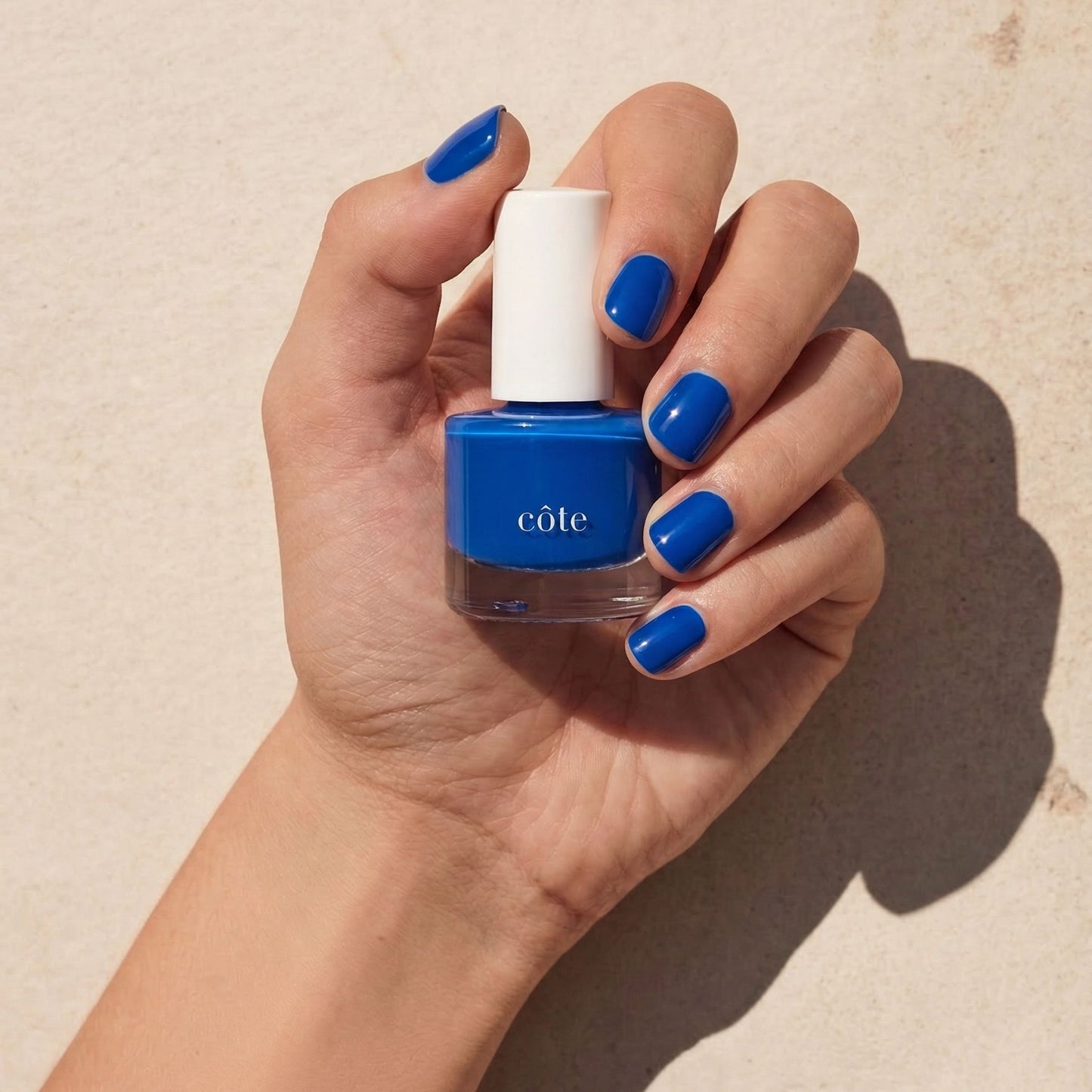 Hand holding Côte Beauty Blue Jeans true blue non-toxic nail polish bottle with nail painted to match.
