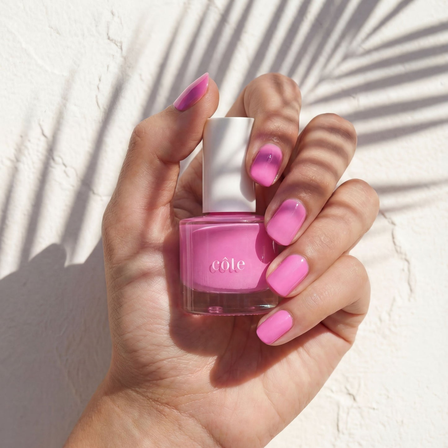 Hands with nails painted holding Côte Beauty Blush & Bashful rich rosy pink non-toxic nail polish bottle