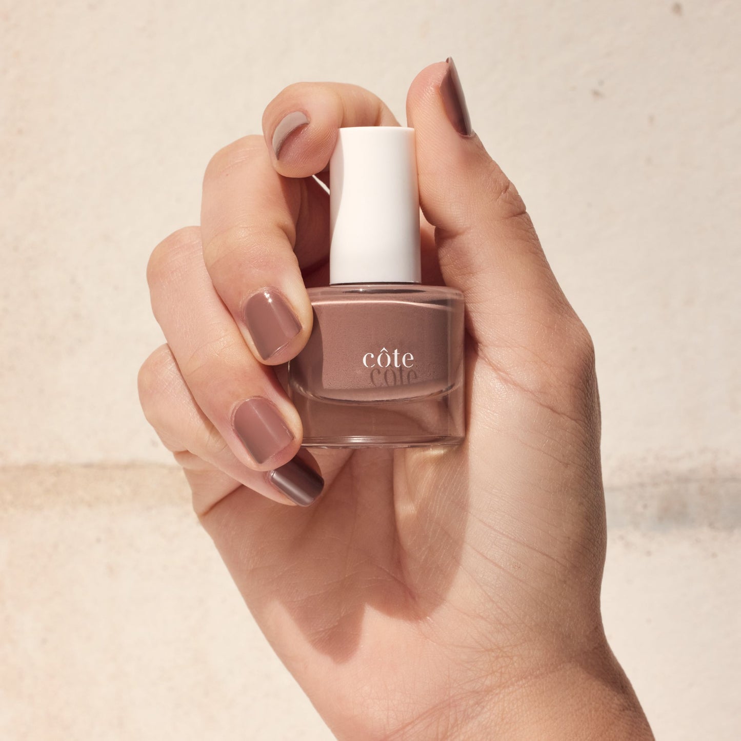 Hand Holding Côte Beauty Cappuccino taupe brown non-toxic nail polish bottle with nails painted to match.