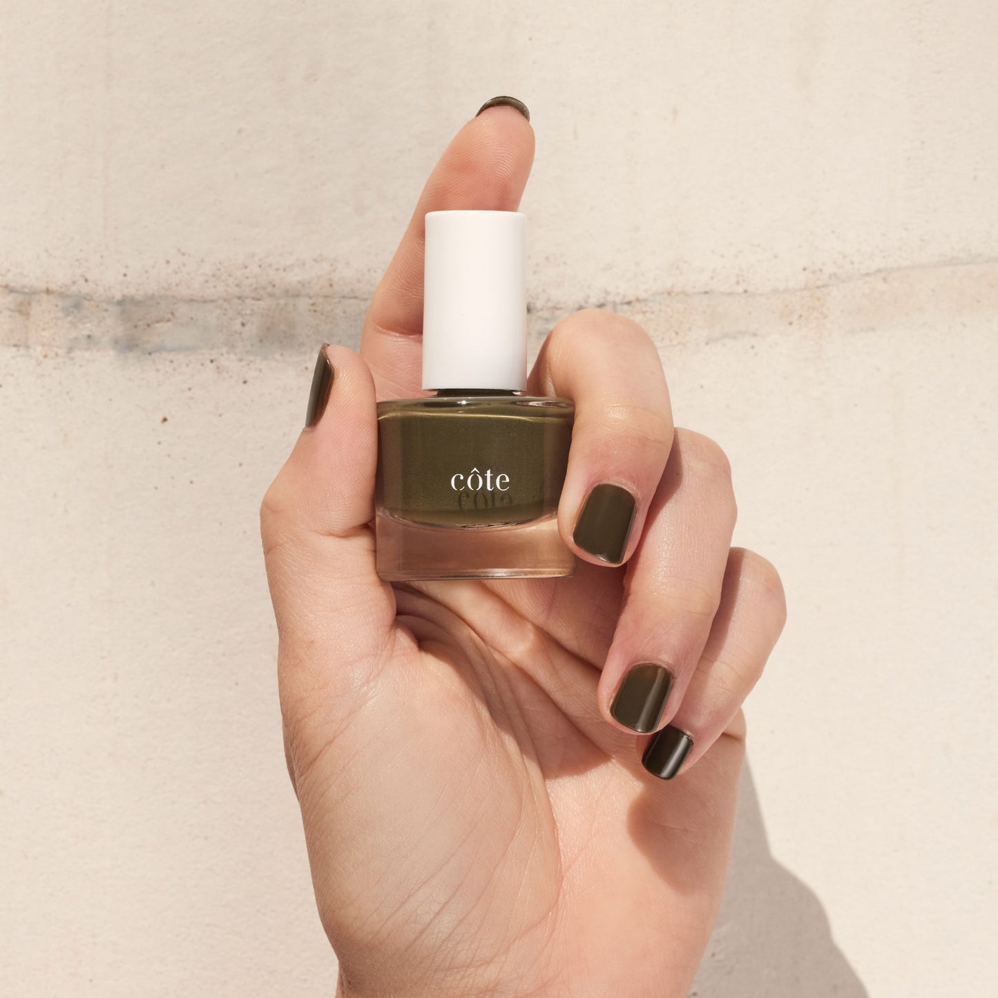 Hand holding Côte Beauty Smokey Sage earthy olive-brown non-toxic nail polish bottle with nails painted to match.