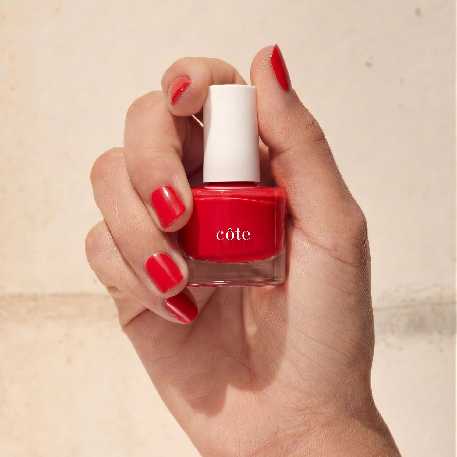 Hand holding Côte Beauty Summer of Love Vibrant Classic Red Non-Toxic Nail Polish Bottle with nails painted to match.