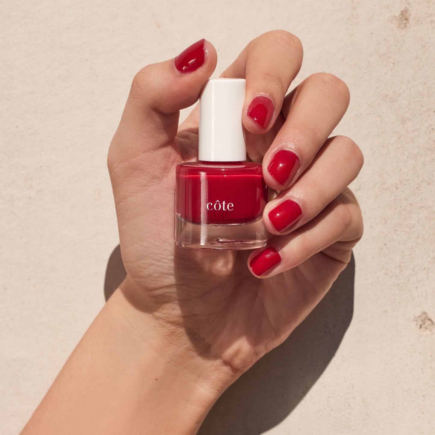 Hand holding Côte Beauty Tango Deep Classic Red Non-Toxic Nail Polish with nails painted to match. 
