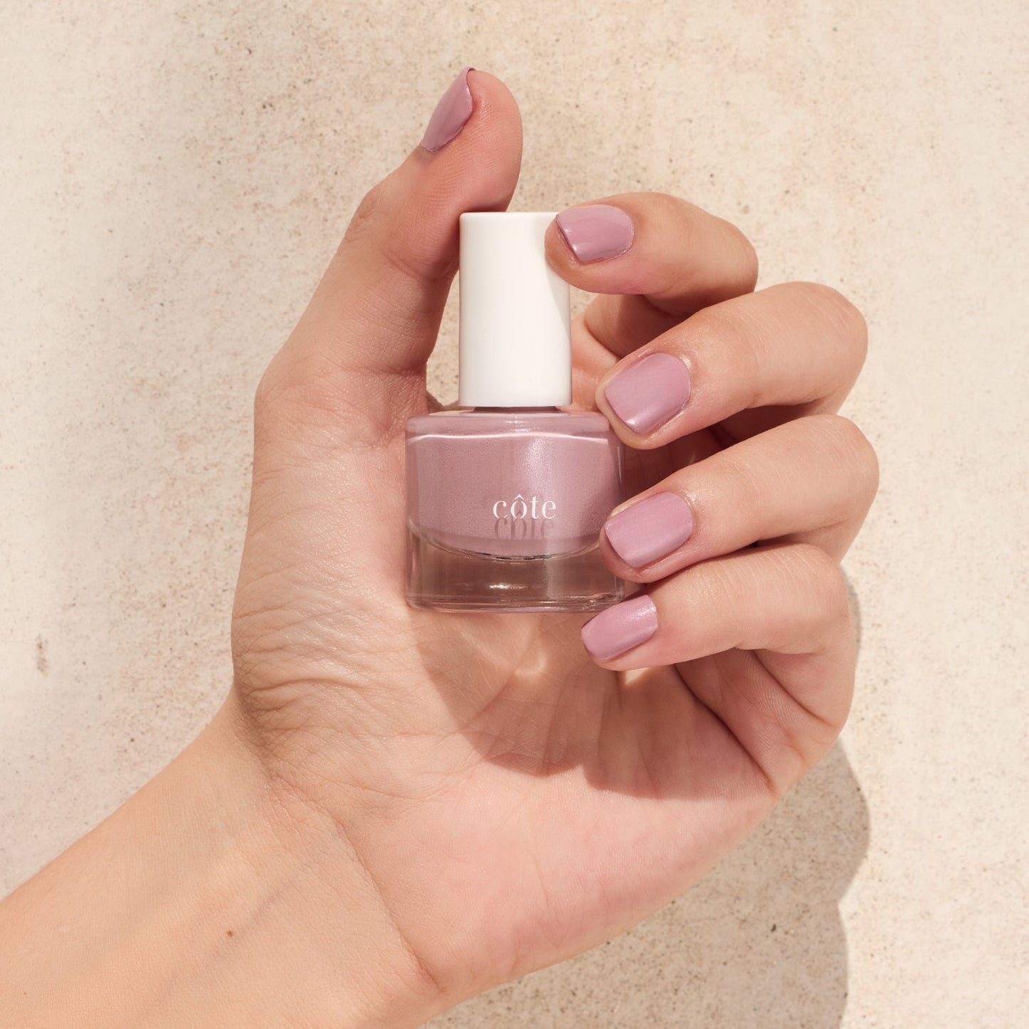 Hand holding nail polish bottle with nails painted with Côte Beauty Wistful shimmering wisteria mauve non-toxic nail polish.